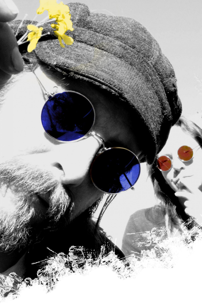 Portrait mit Mütze und blauer Brille, im Hintergrund eine Frau mit roter Brille